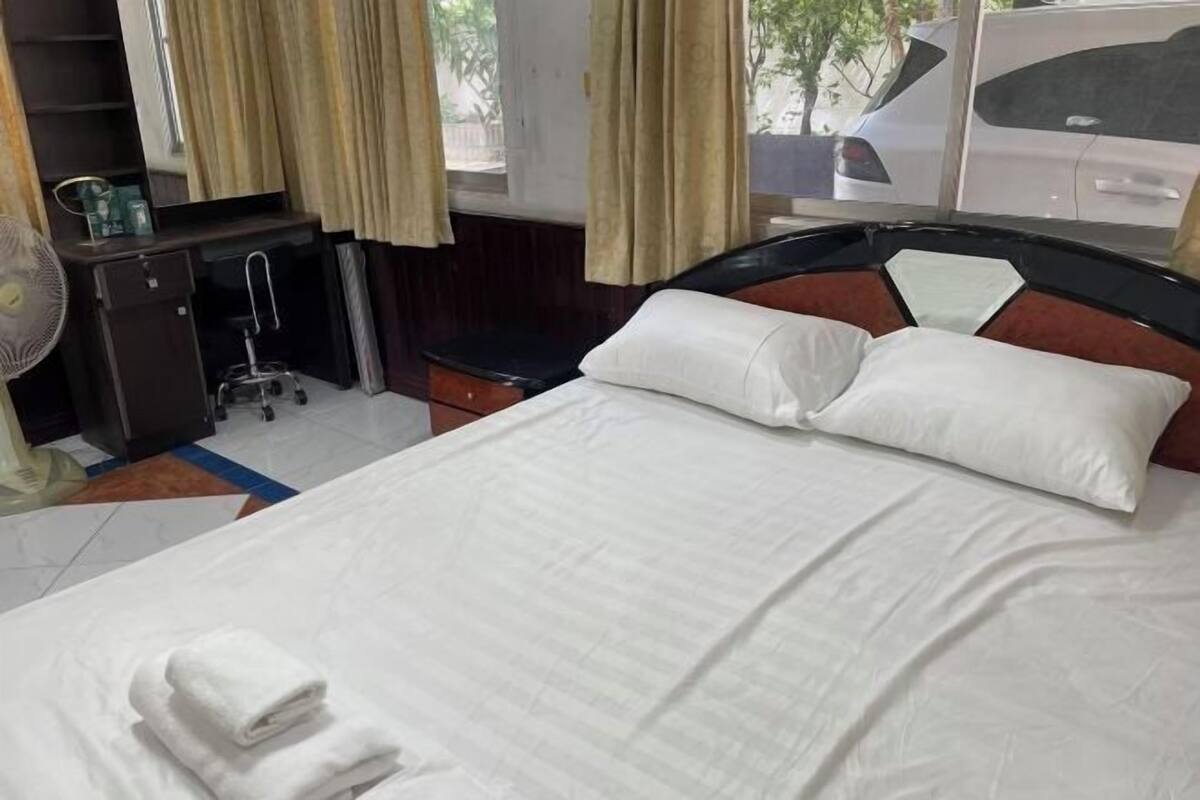 A hotel room with a bed, a desk with a mirror, a fan, and a window with curtains.
