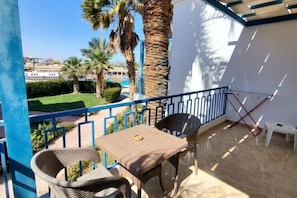 A balcony with a table and chairs, a white wall, and a view of a green garden with palm trees. - HRG Hurghada Aparthotel Aqua park A100 - By MAMO Property (Hurghada)