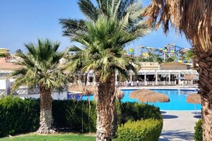 A pool area with palm trees, a clear blue pool, and a distant amusement park. - HRG Hurghada Aparthotel Aqua park A100 - By MAMO Property (Hurghada)