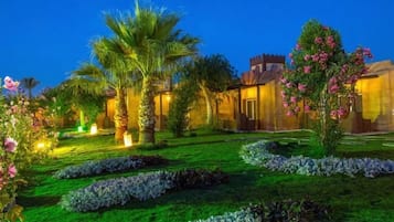 A well-maintained garden with palm trees and flowering plants, a building with a traditional architectural style, and a clear sky.