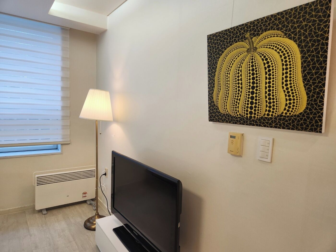 A modern room with a flat-screen TV, a standing lamp, a radiator, and a wall-mounted artwork.