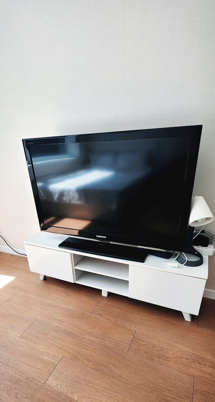 Litteänäyttöinen televisio on sijoitettu valkoiselle TV-tasolle, ja sen vieressä on kaukosäädin ja lamppu.