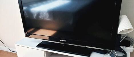 Litteänäyttöinen televisio on sijoitettu valkoiselle TV-tasolle, ja sen vieressä on kaukosäädin ja lamppu.