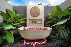 A freestanding bathtub with a decorative flower arrangement, surrounded by lush green plants and a stone pathway. - Villa Anahata by BaliSuperHost (Bali)