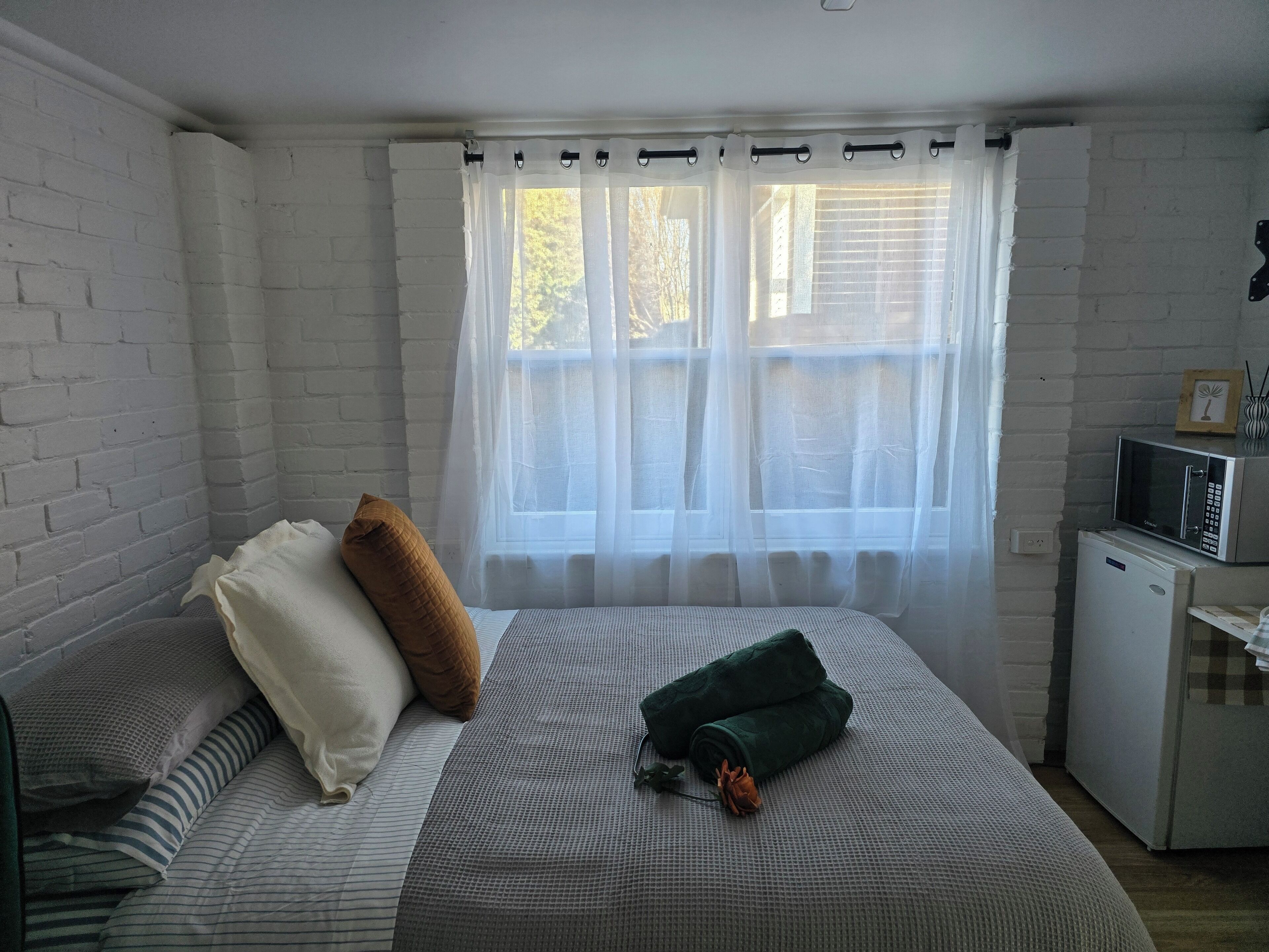 Une chambre à coucher avec un lit, des oreillers, une table de chevet avec un micro-ondes, et une fenêtre avec des rideaux légers.