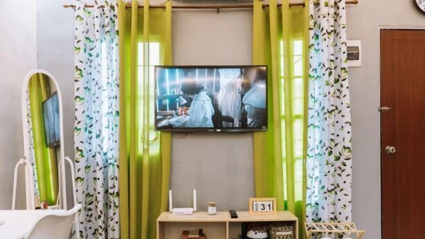 Ein Zimmer mit an der Wand montiertem Fernseher, einer kleinen Holzablage mit Büchern und einer Uhr sowie einem Schuhregal mit Hausschuhen.