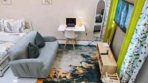 Ein Schlafzimmer mit grauem Sofa, einem Schreibtisch mit Laptop, einem Bett mit gemusterten Betttextilien und einem farbenfrohen Teppich.