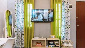 A room with a television mounted on the wall, a small wooden shelf with books and a clock, and a door with a number sign.