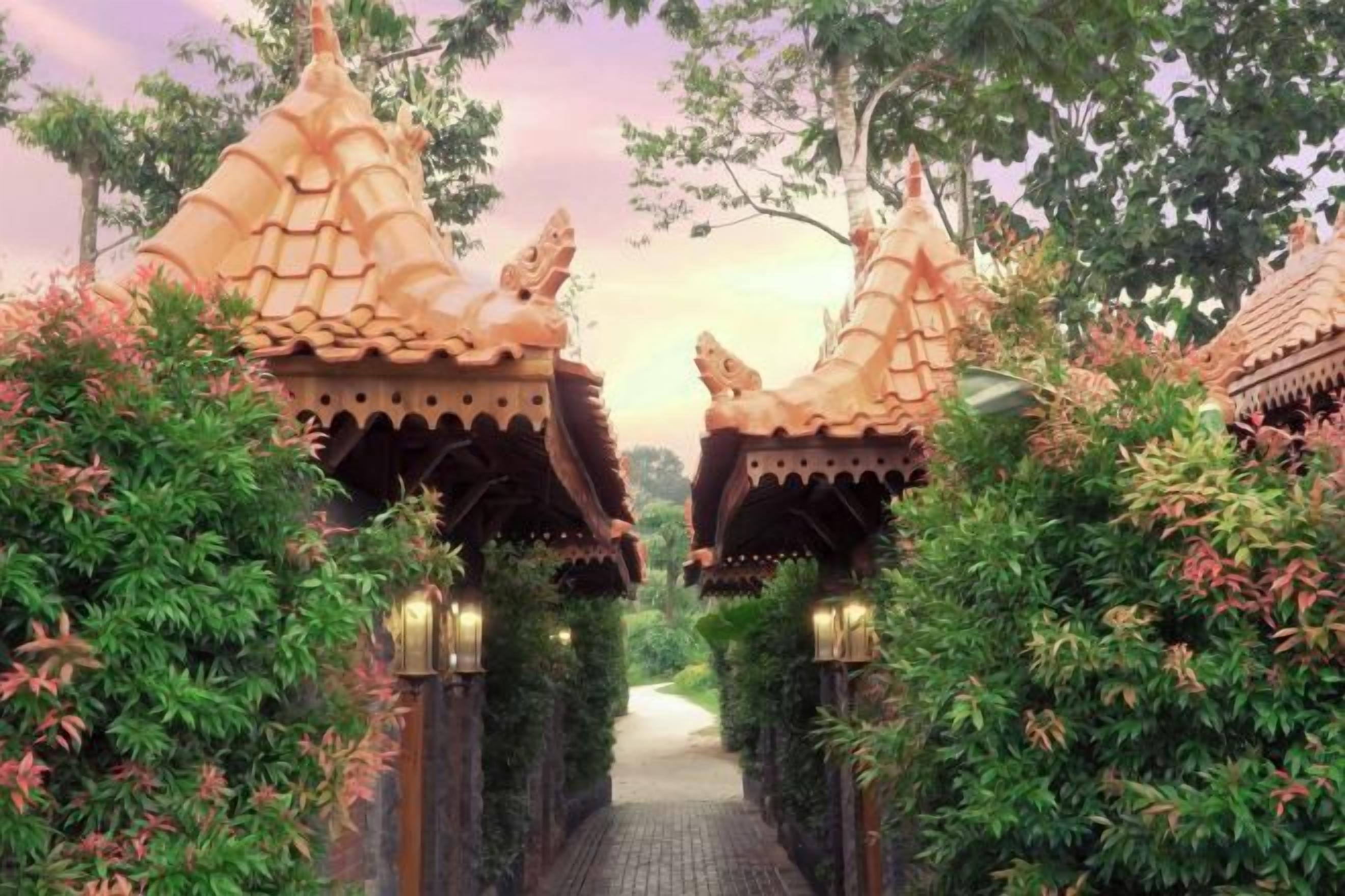 Un sentier mène à des bâtiments de style traditionnel, dotés de toits richement décorés, et entourés d’une végétation luxuriante.