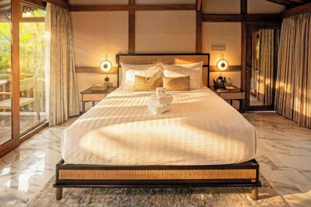 Un grand lit avec un cadre en bois, deux tables de chevet avec des lampes et une vue dégagée sur l’extérieur grâce à une grande fenêtre.