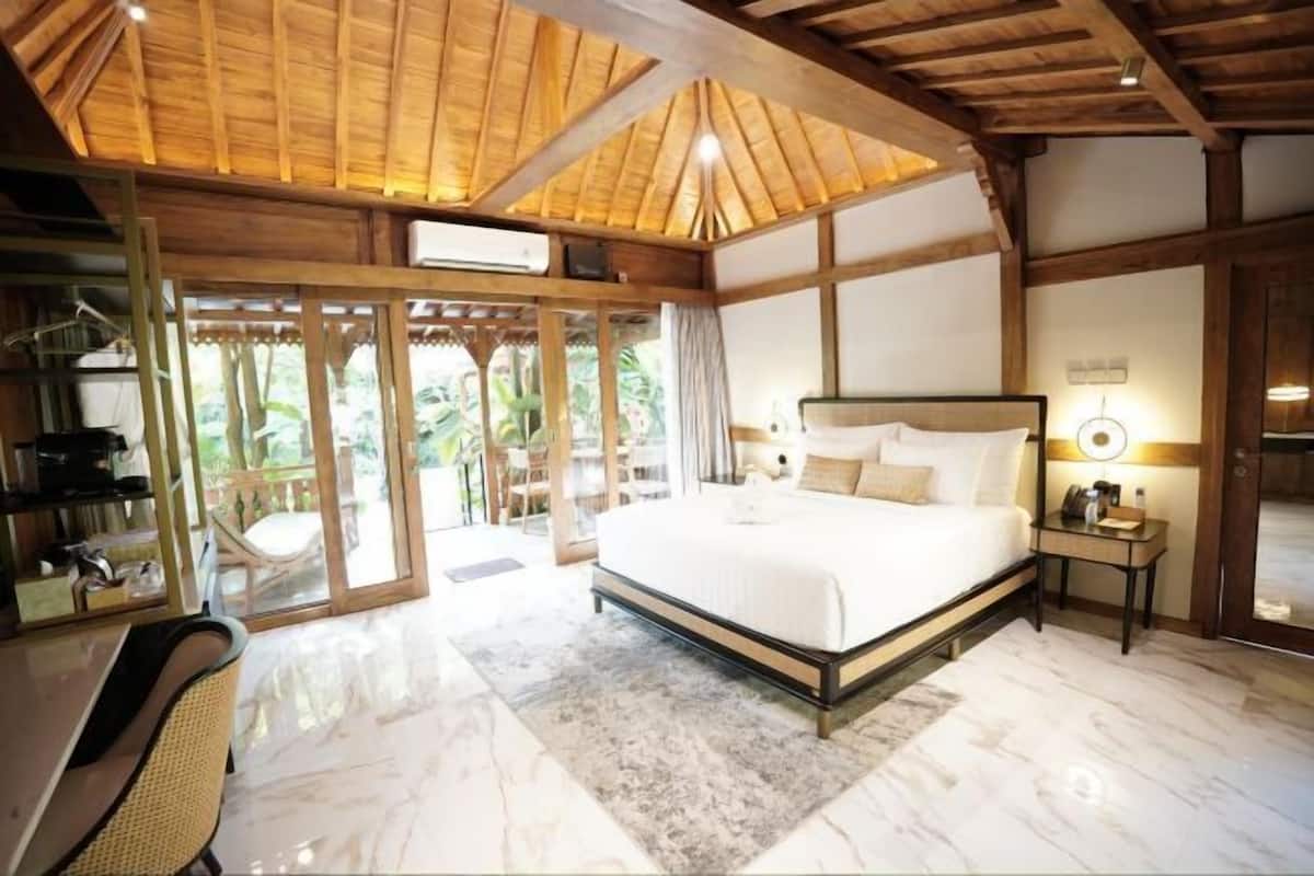 Une chambre spacieuse avec un grand lit, des poutres apparentes en bois au plafond, un bureau avec une chaise et une vue sur la verdure grâce à des portes vitrées.