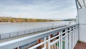 Une vue sur la rivière depuis un balcon doté d’une balustrade, avec un ciel dégagé et des bâtiments qui se dressent au loin.