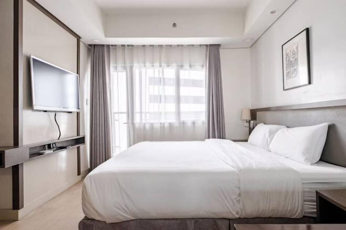 A hotel room with a bed, a TV mounted on the wall, a window with curtains, and a framed picture on the wall.