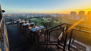 A rooftop terrace with tables and chairs, offering a view of a cityscape and a park during sunset. - Cozy 1 Studio - The Residences BGC Taguig (Taguig)