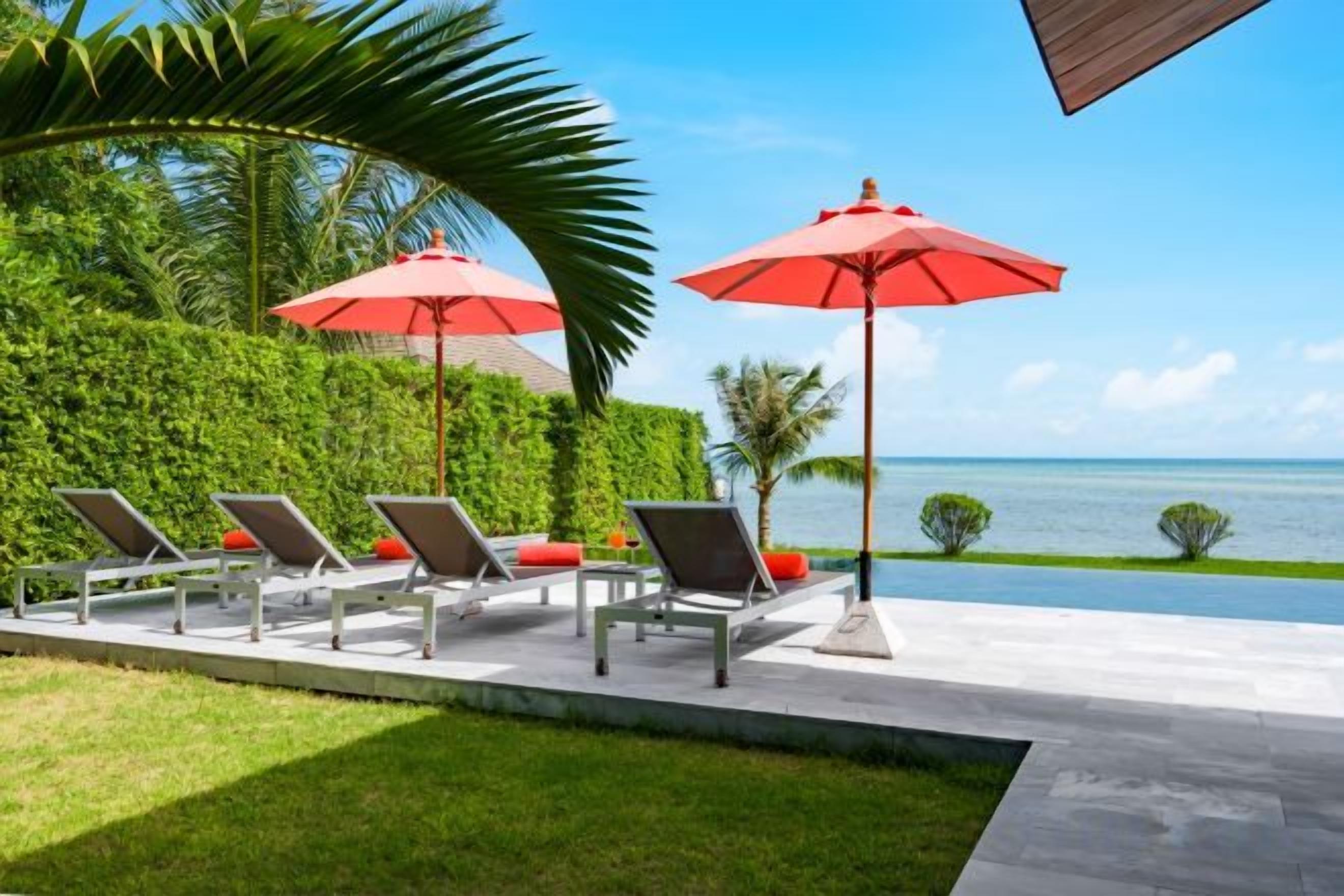 Zona junto a la piscina con sillones de descanso y sombrillas rojas, con vistas a una masa de agua.