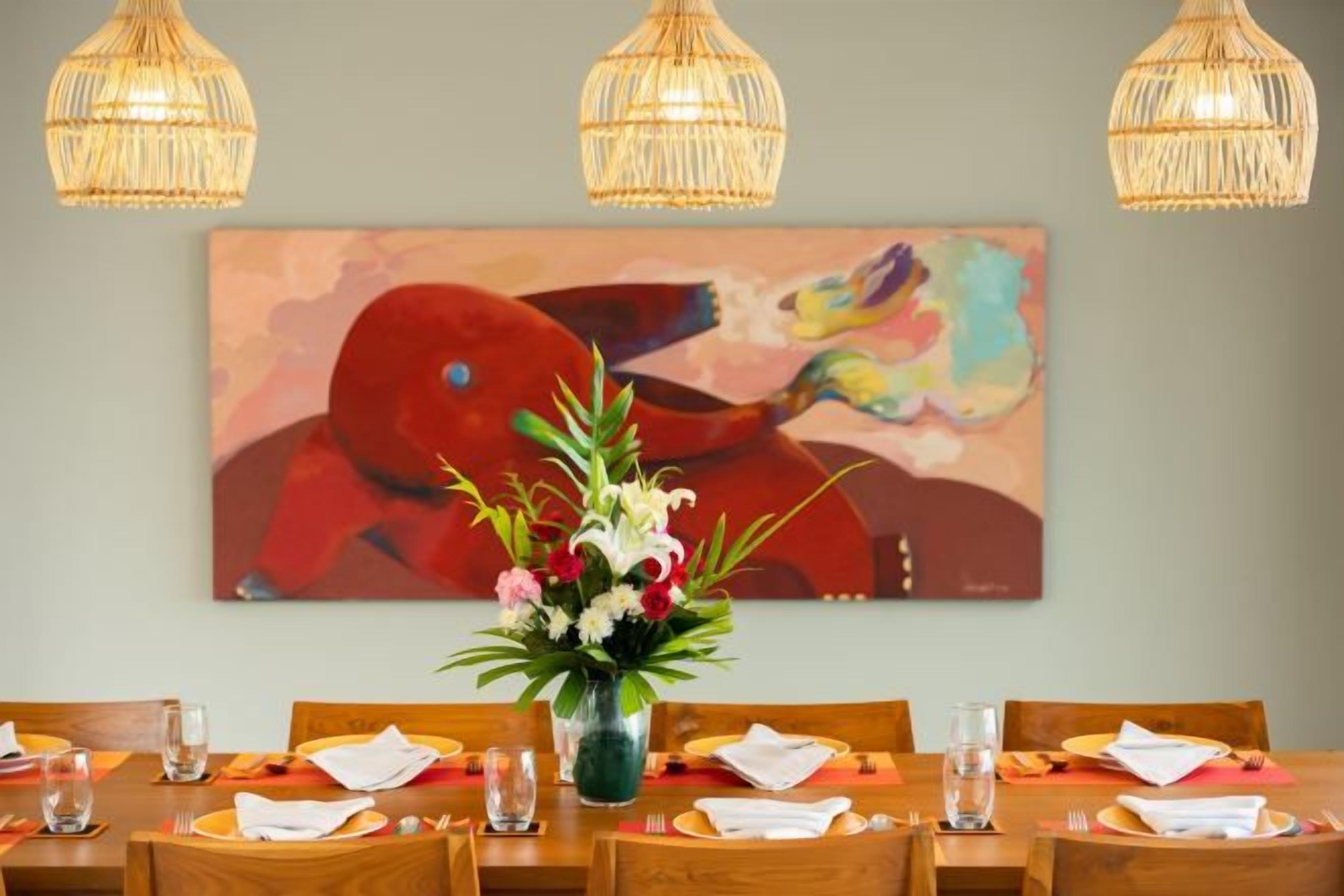 Un comedor con una mesa preparada para cuatro, un florero con flores y un cuadro abstracto y colorido en la pared.