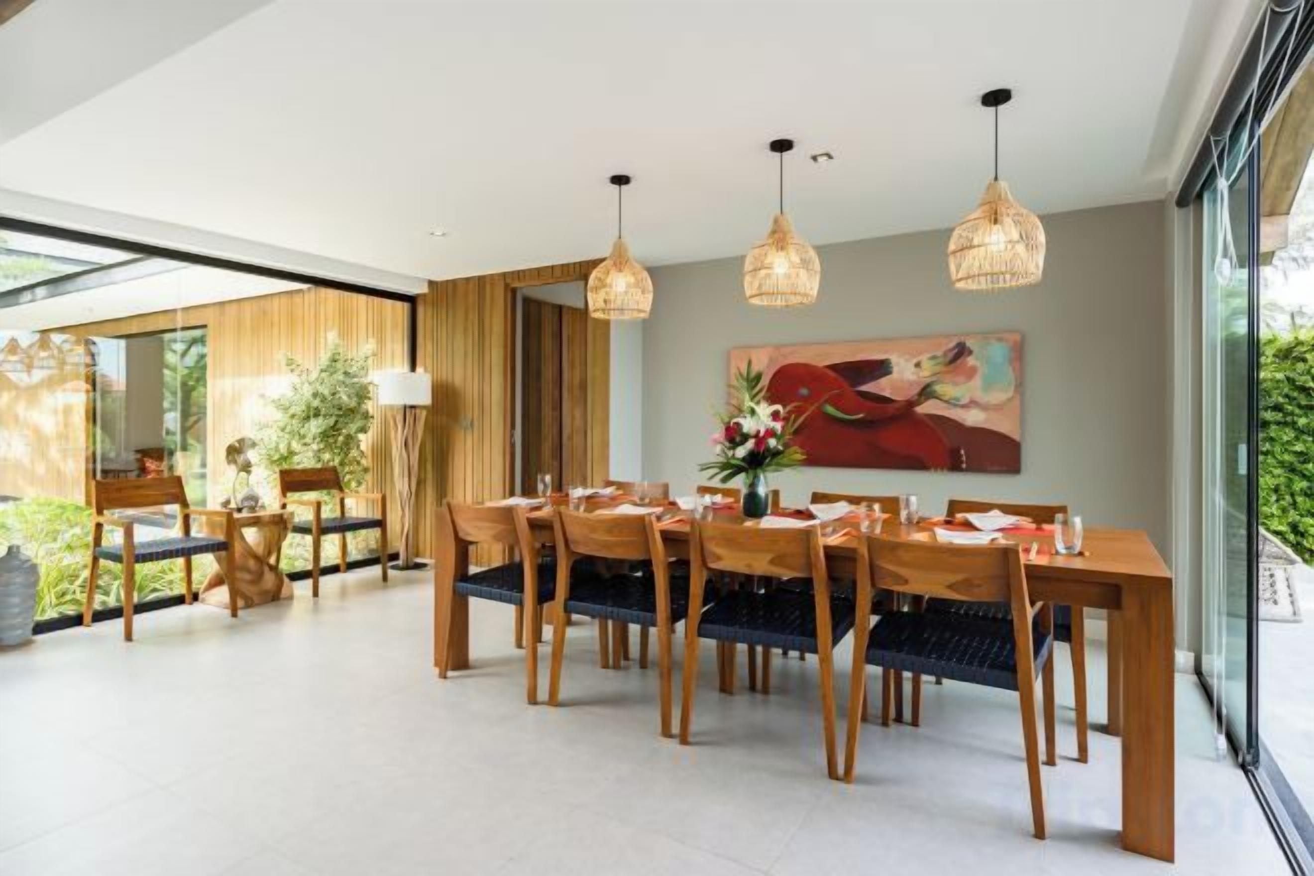 Un comedor con una larga mesa de madera, varias sillas, lámparas colgantes y un cuadro grande en la pared.