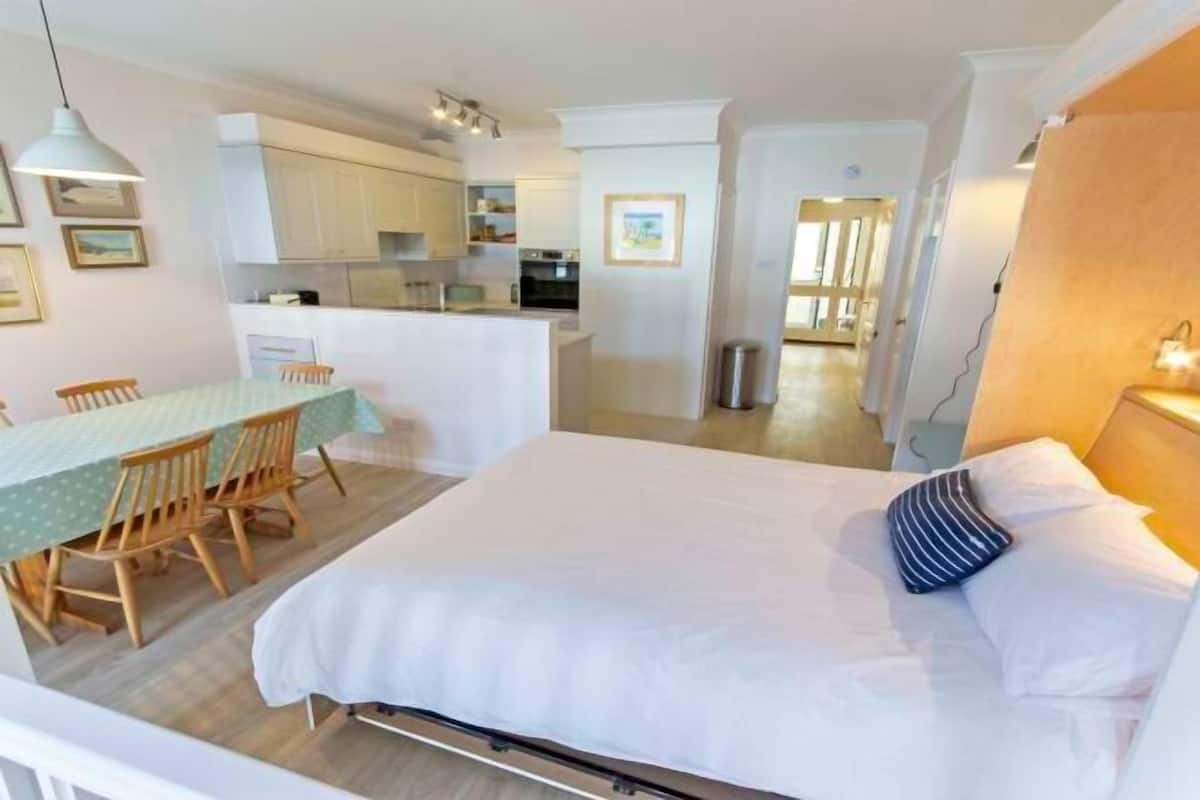 A bedroom with a bed, bedside table, and a view into the kitchen.