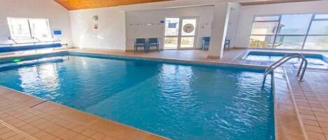 Una piscina interior con suelo de baldosas, techo de madera y vistas al exterior a través de amplias ventanas.