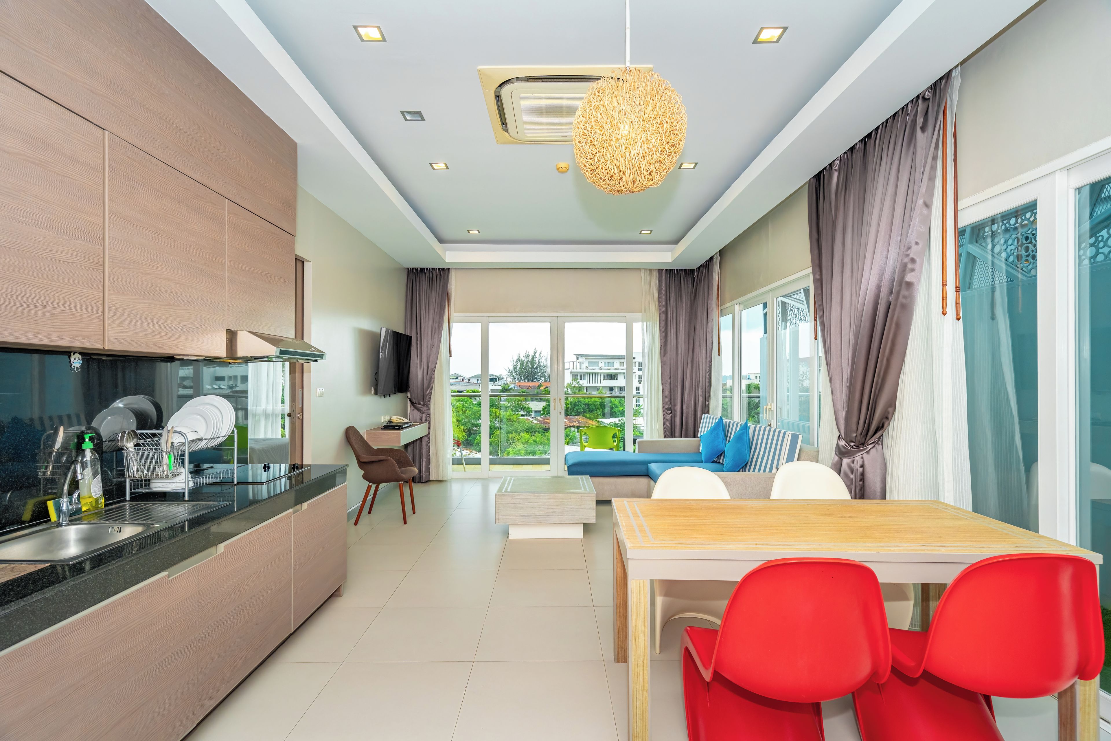 A modern kitchen with a dining area, a sofa, and a view of greenery outside.