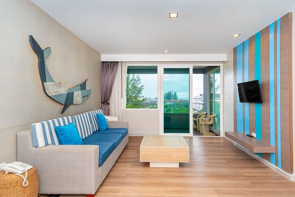 A modern living room with a sofa, a coffee table, a mounted artwork, a television, and a sliding glass door leading to an outdoor area. - Ratana Platinum Resort Kamala (Kamala)