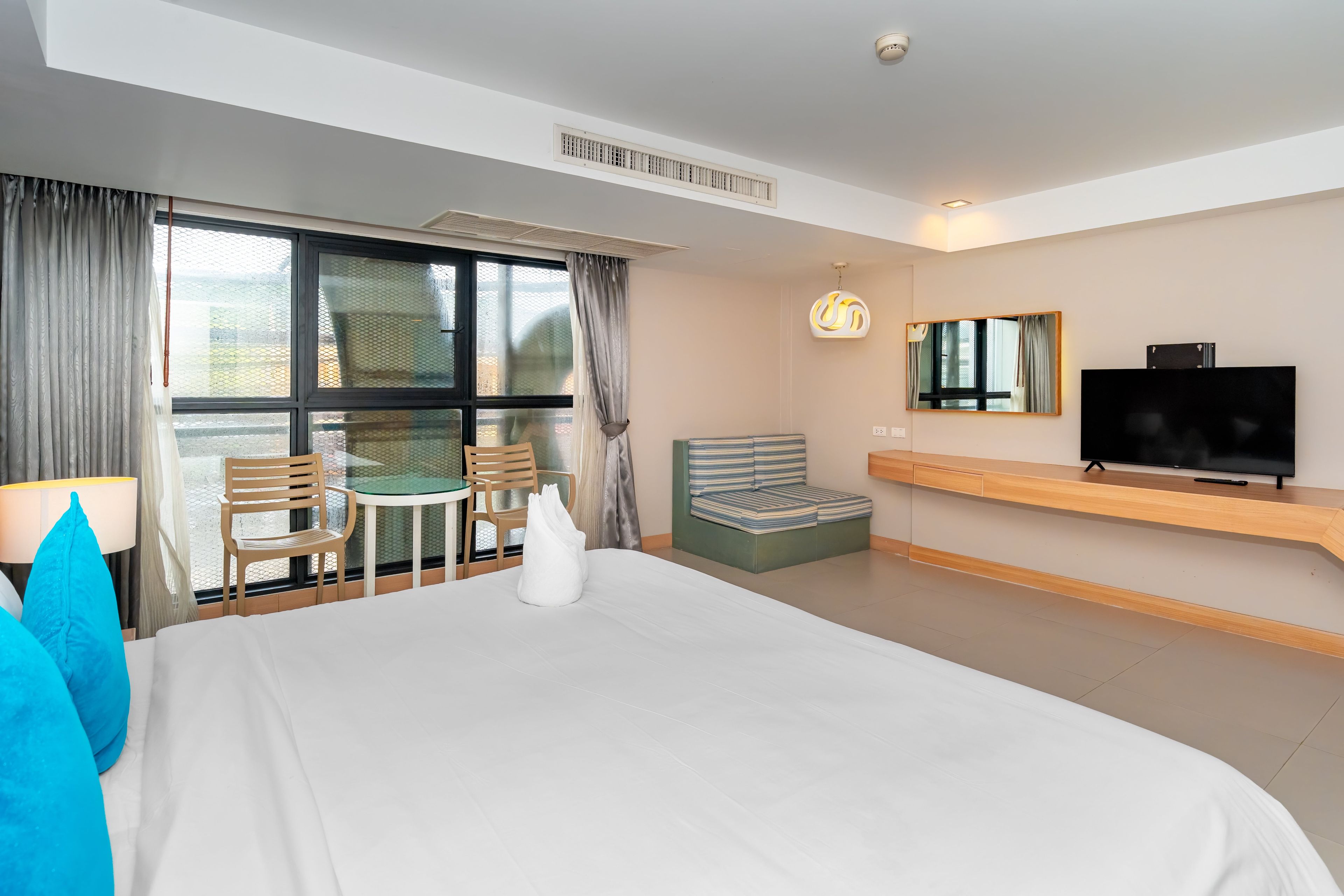 Quarto de hotel com uma cama grande, uma televisão, área de estar com mesa e cadeiras, e varanda com vista.
