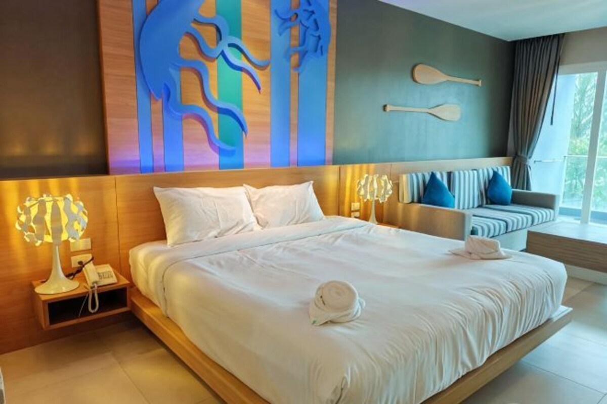A modern hotel room with a large bed, a bedside table with a lamp, a seating area with cushions, and a view of the outdoors.