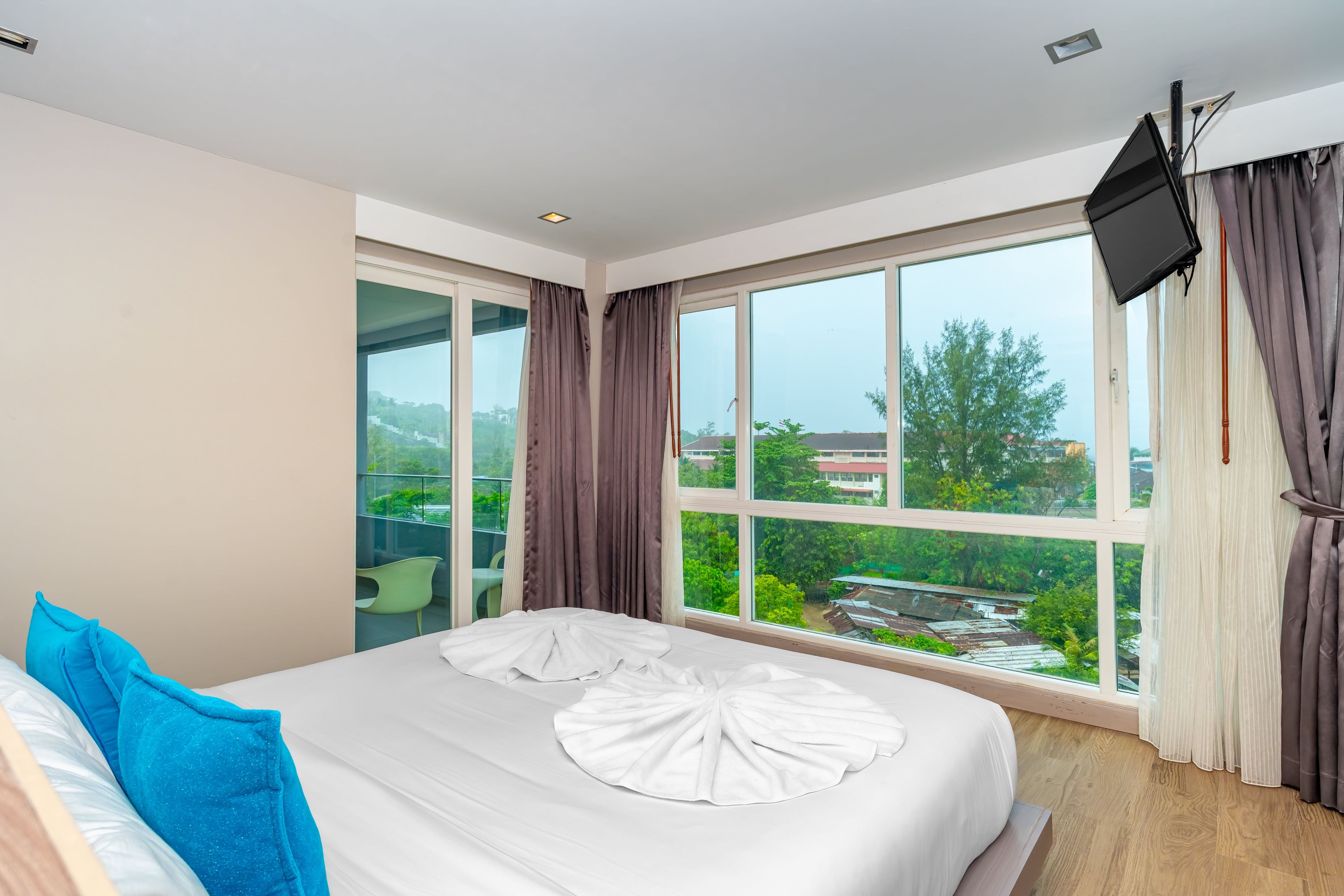 A hotel room with a large window, a bed with white bedding, a blue pillow, and a flat-screen TV mounted on the wall.