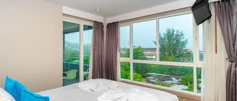 A hotel room with a large window, a bed with white bedding, a blue pillow, and a flat-screen TV mounted on the wall.