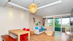 Een moderne woonkamer met een houten vloer, een eettafel met rode stoelen, een blauwe bank met kussens en een groot raam met uitzicht op groen.