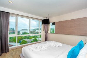Habitación de hotel con un ventanal grande, cama con ropa blanca, televisor de pantalla plana y vistas a edificios y árboles.