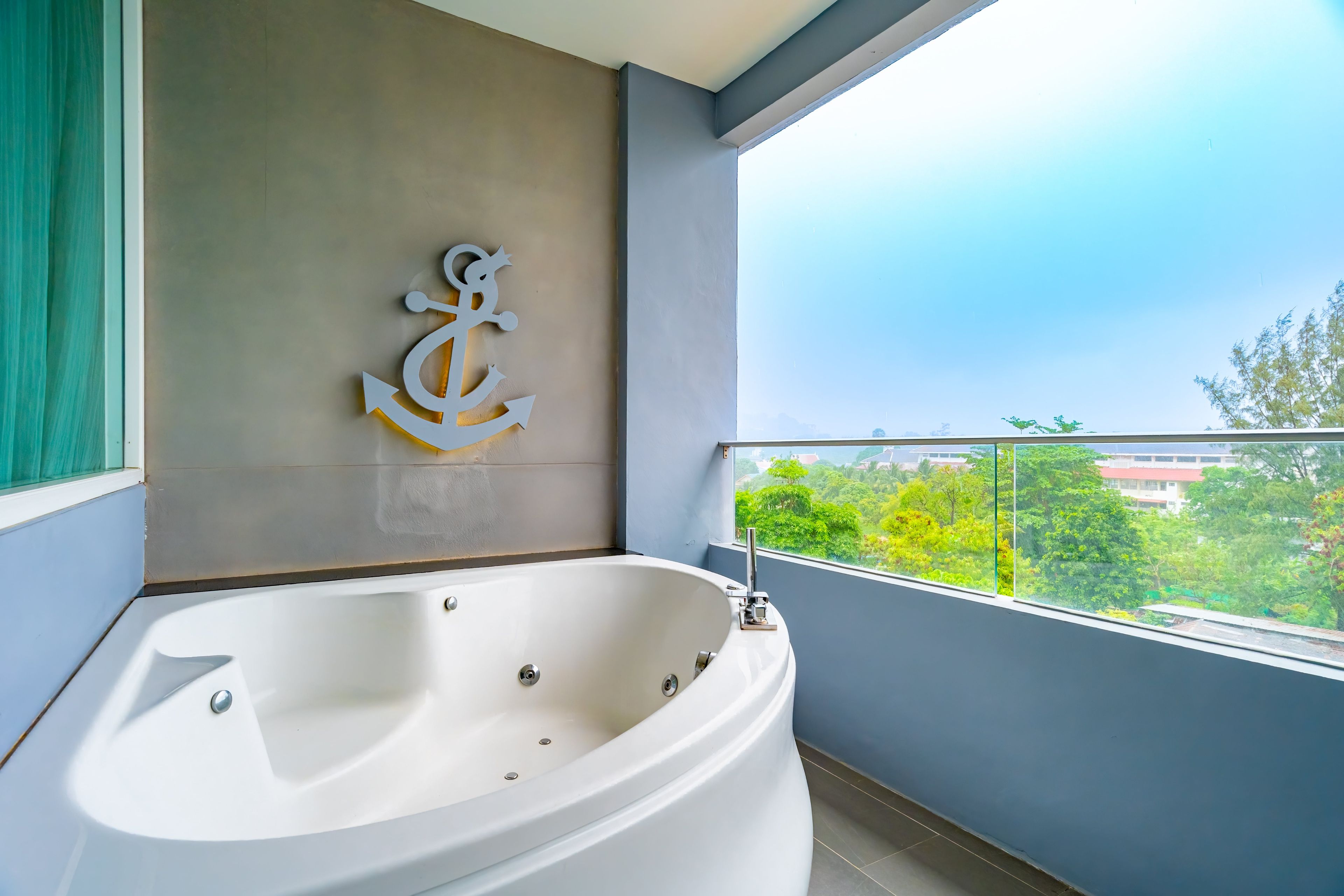 A modern bathroom with a large bathtub, a decorative anchor wall art, and a balcony with a view of greenery.