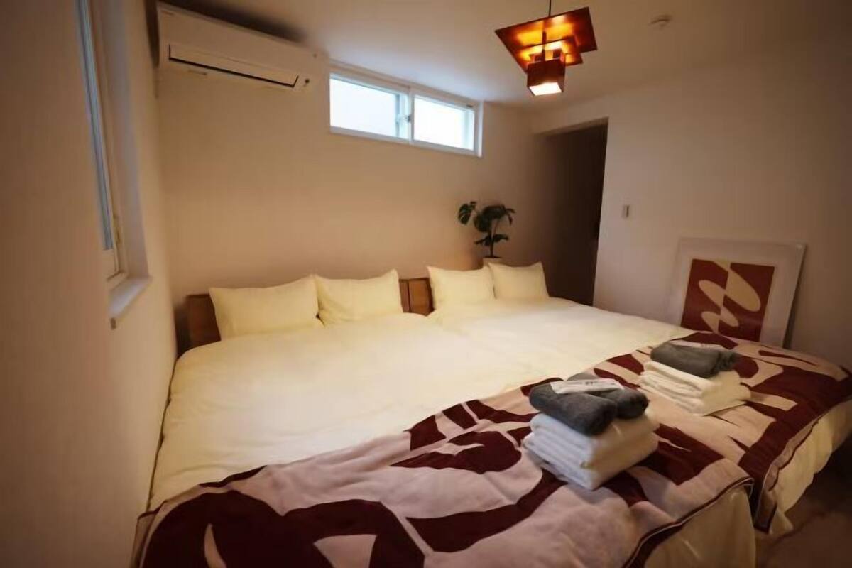 A bedroom with a large bed, a window, an air conditioning unit, and a wall-mounted light fixture.
