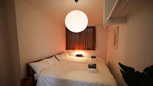 A double bed with a white bedspread, a bedside table with a lamp, a large potted plant, and a window with curtains. - URBAN HOUSE KOENJI (Tokyo)