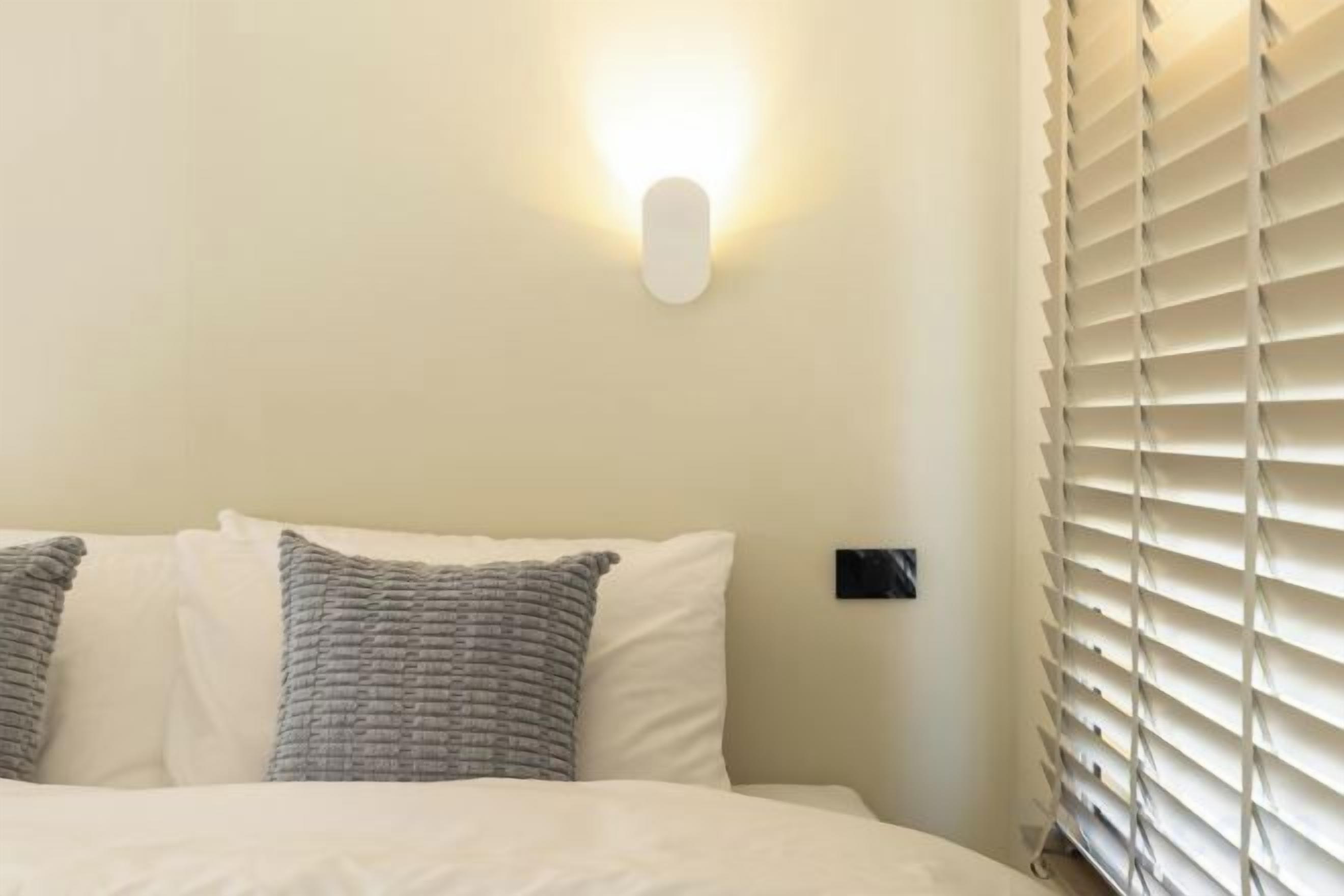 A bed with white bedding and two pillows, a wall-mounted light fixture, and a window with blinds.