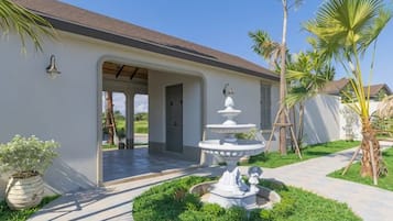 A house with a covered porch, a white fountain, and a well-maintained garden with palm trees.