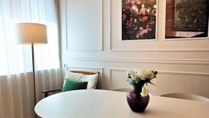 Een kamer met een ronde tafel, een vaas met bloemen, een lamp en lijstjes met kunstwerken aan de muur.