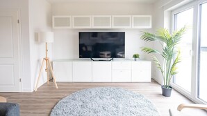 Ein modernes Wohnzimmer mit einem Flachbildfernseher, weißen Schränken, einem runden grauen Teppich, Holzboden und einem großen Fenster mit einer Pflanze.