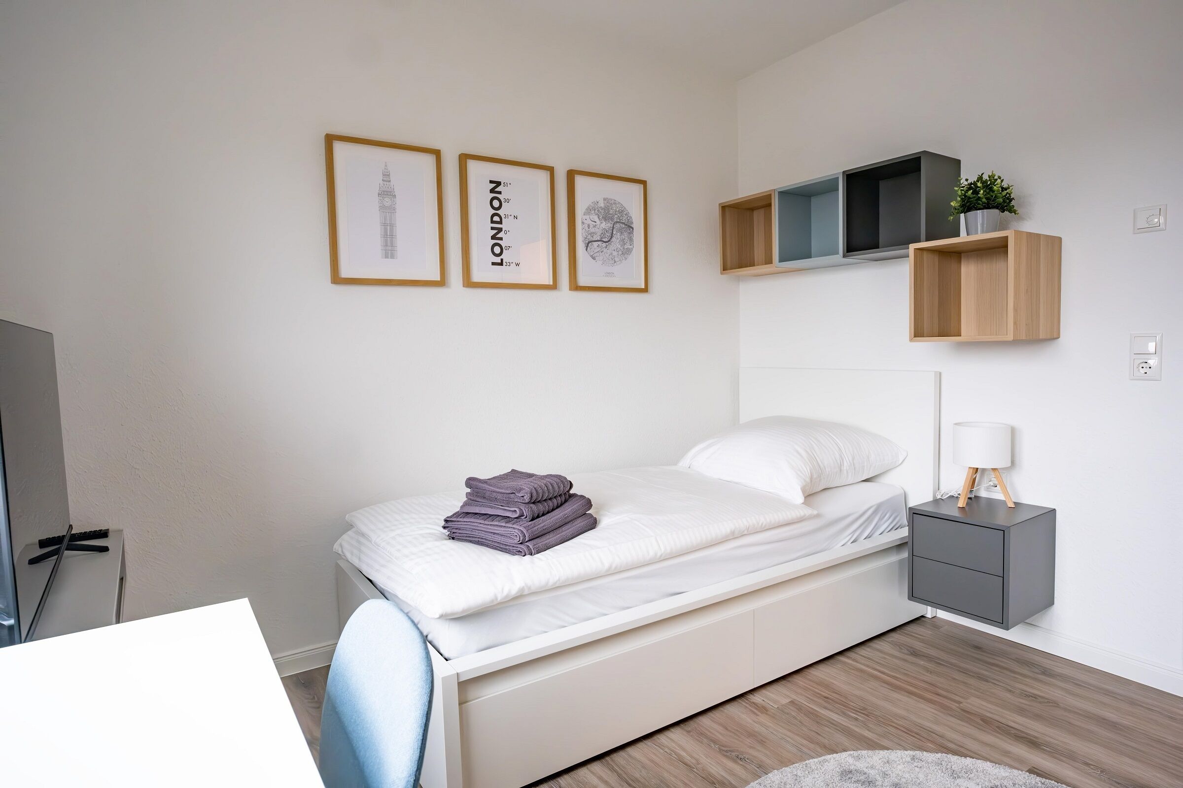 Une chambre à coucher comprenant un lit, une table de chevet, une lampe, un téléviseur et des étagères fixées au mur.