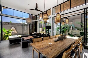 A modern dining area with a wooden table and chairs, a sofa, and hanging pendant lights. - Villa Swing by BaliSuperHost (Bali)