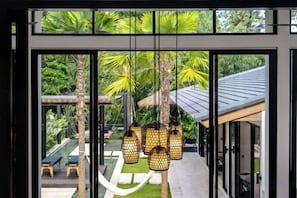 A view through a glass door into a tropical outdoor area with hanging lanterns, palm trees, and a hammock. - Villa Swing by BaliSuperHost (Bali)
