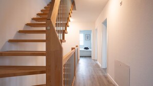 A wooden staircase with a handrail leading to a landing. A hallway with a light switch and a door is visible in the background.
