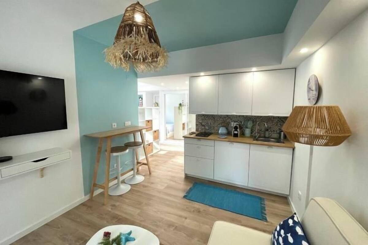 Un salon moderne avec un téléviseur, un coin repas avec une table et des chaises, et une cuisine avec des meubles blancs et un tapis bleu.