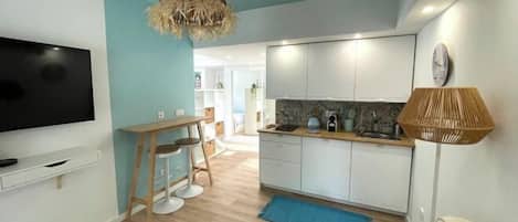 A modern living room with a TV, a dining area with a table and chairs, and a kitchen with white cabinets and a blue rug.