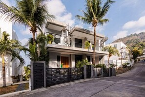 Una casa de dos pisos con un balcón, rodeada por una cerca negra, palmeras y un cielo despejado.