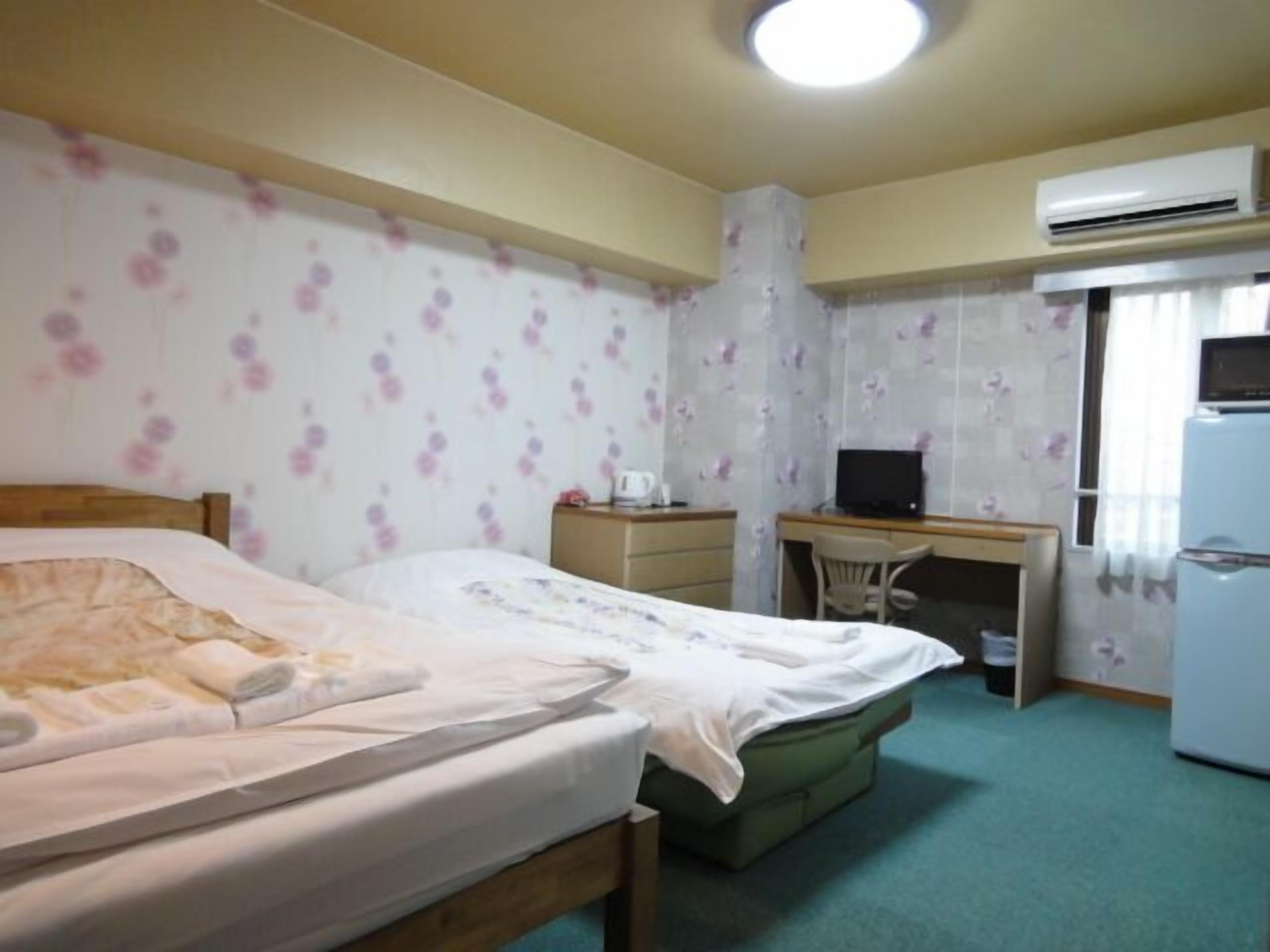 A hotel room with a bed, a desk, a chair, an air conditioner, and a refrigerator.