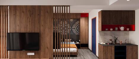 A modern hotel room with a wooden partition, a flat-screen TV, a bed, a desk, and a kitchen area with cabinets and a sink.
