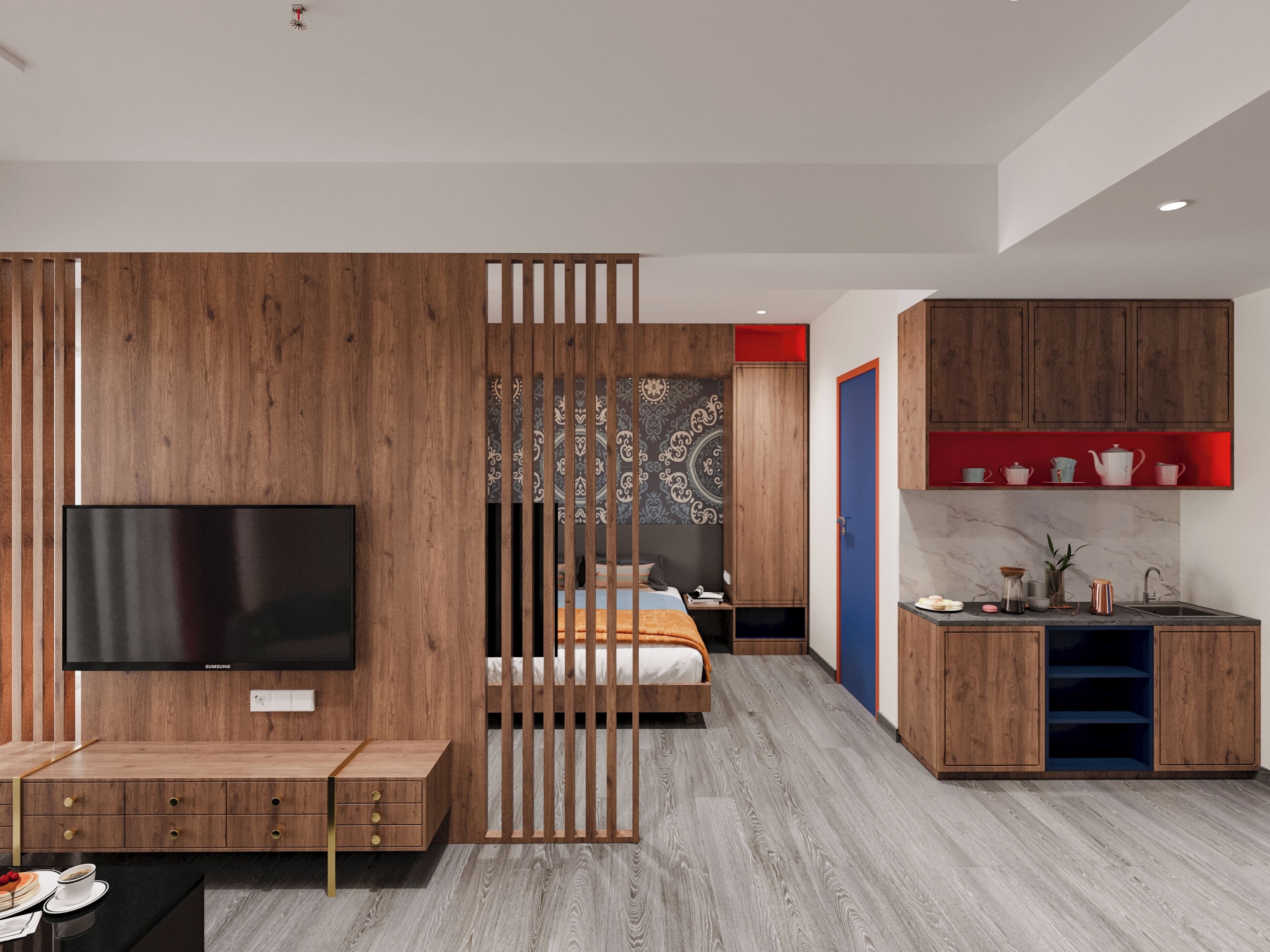 A modern hotel room with a wooden partition, a flat-screen TV, a bed, a desk with a sink, and a red shelf with kitchenware.