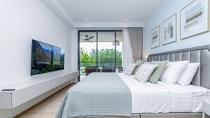 A modern bedroom with a large bed, a flat-screen TV, and a balcony with a view of greenery. - Beachside 2BR 200 m to BangTao beach (Choeng Thale)