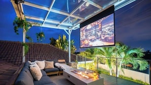 An outdoor living area with a modern sofa, a coffee table with a fire feature, and a large screen displaying a cityscape. - Kira Japandi Escape with Sauna & Rooftop cinema (Hua Hin)
